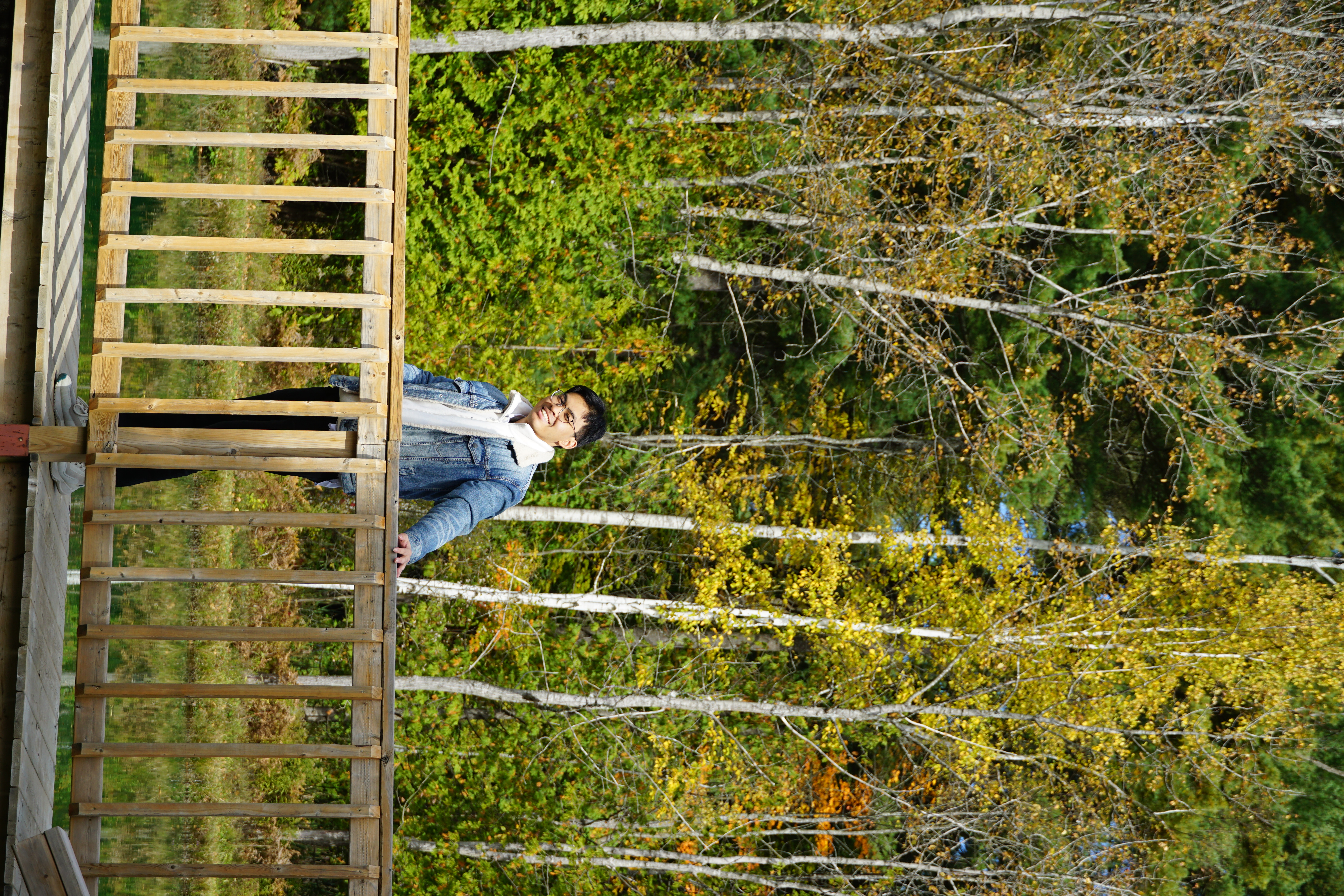 A young man standing on a wooden bridge in a forested area. He is smiling, wearing a denim jacket over a hoodie, with tall trees and autumn leaves in the background.