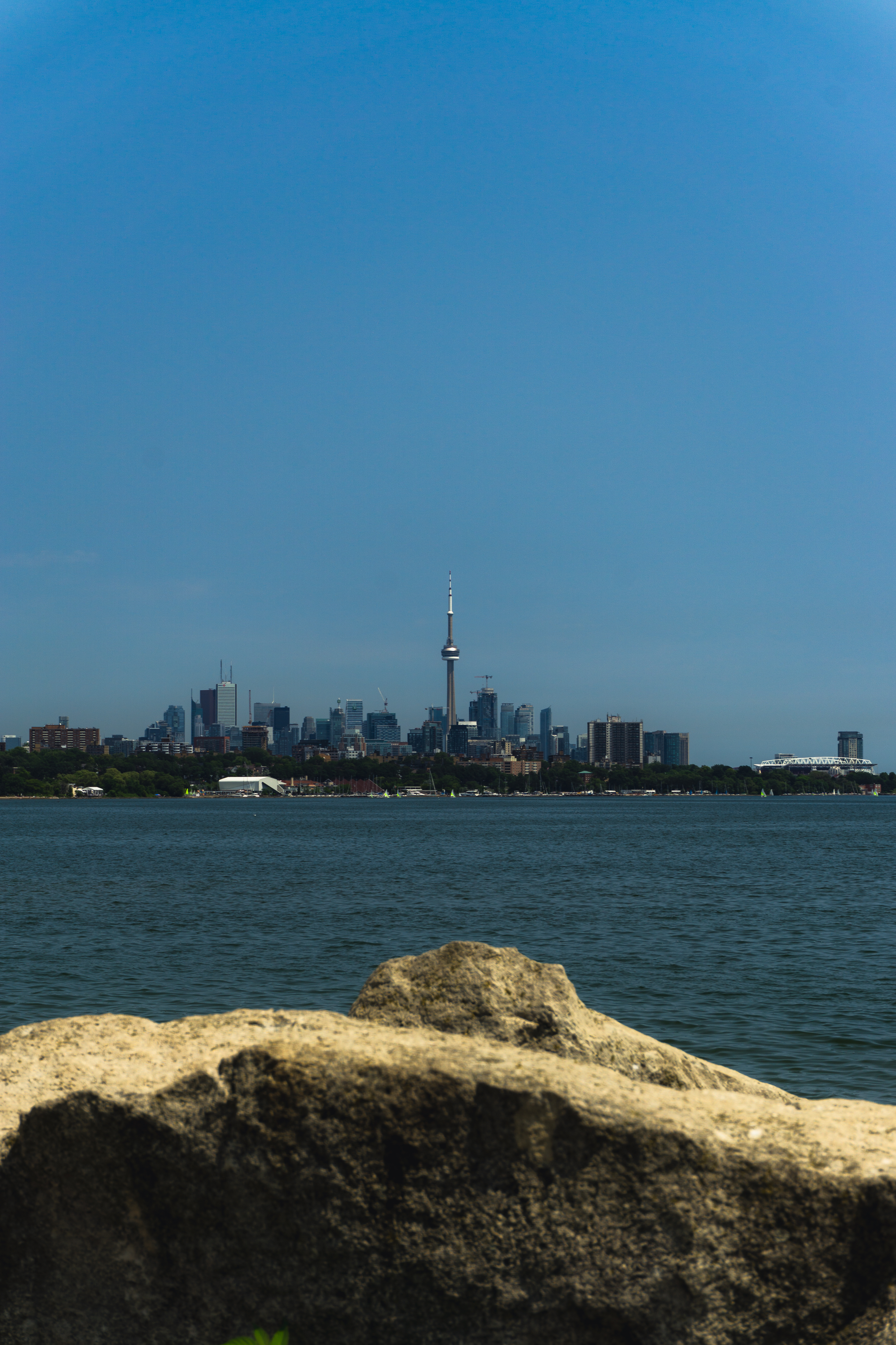 Toronto skyline viewed from across Lake Ontario with large rocks in the foreground. The CN Tower stands tall among the buildings under a clear blue sky on a sunny day.