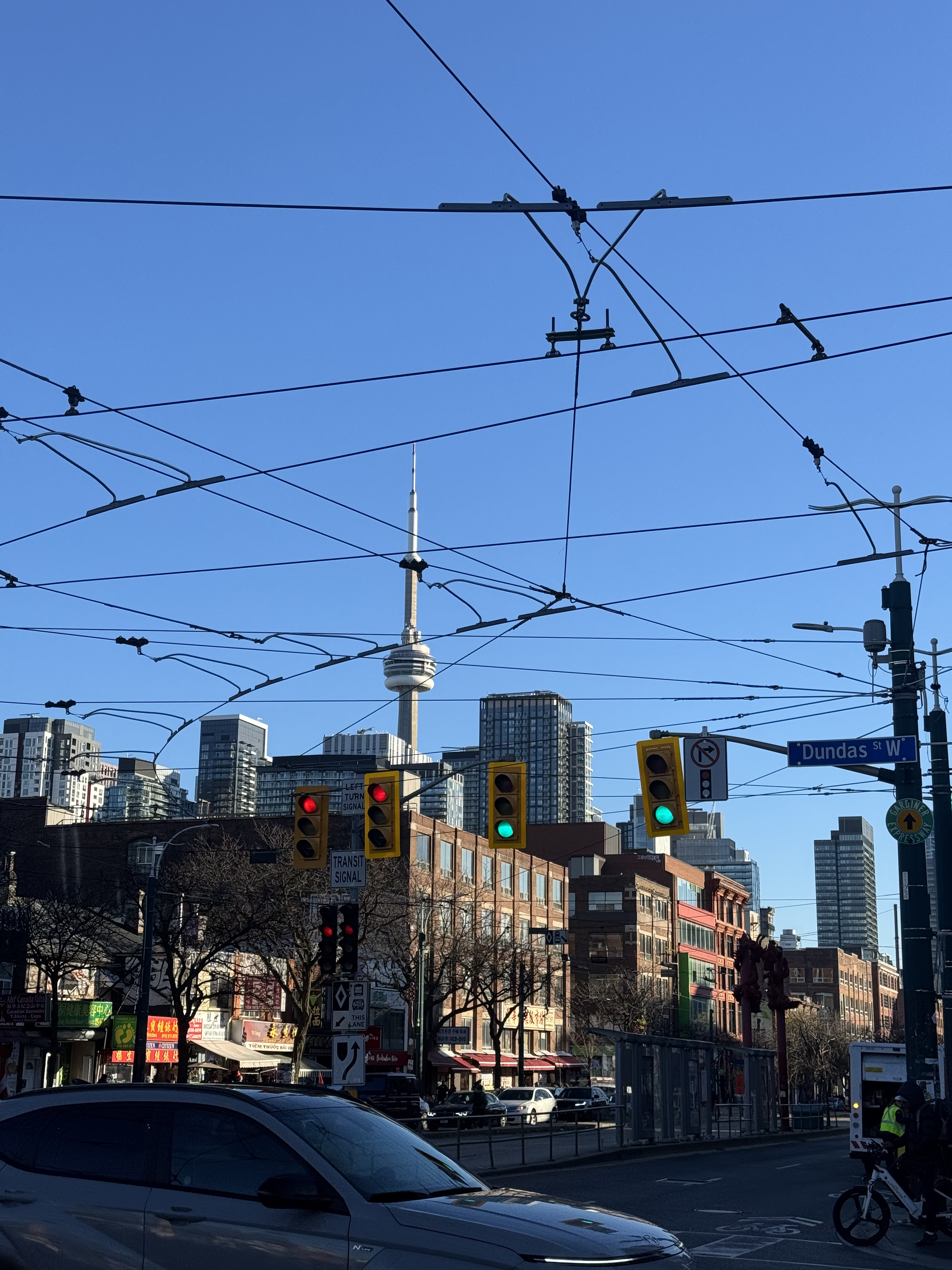 View of Toronto's CN Tower from Spadina Avenue, framed by tall city buildings on either side. The tower stands prominently in the center under a clear sky, capturing the urban feel of downtown Toronto.